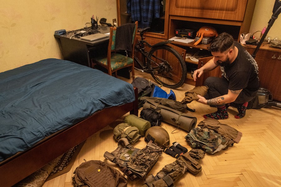 A man kneels down in a room, arranging piles of military gear.