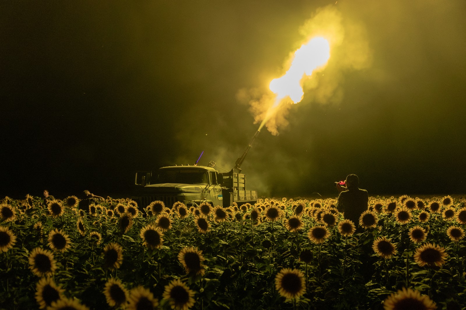 Soldiers fire a large anti-aircraft gun from the back of a truck in a sunflower field, at night, the muzzle flash illuminating the sunflowers.