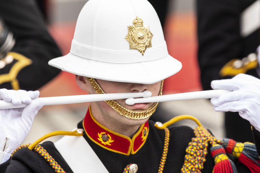 A drummer in a military dress uniform pauses, holding drumsticks up to their mouth.