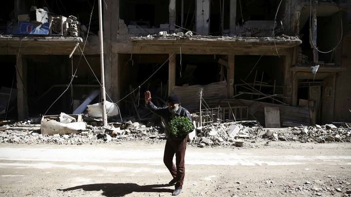 A bearded man waves outside the shell of a bombed-out building.