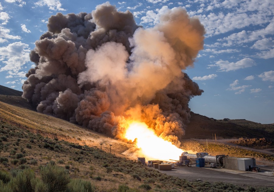 A rocket booster mounted on its side shoots out flame and kicks up a huge cloud of dust during a test.