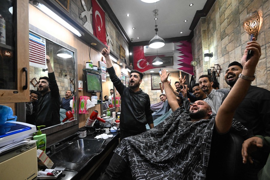 Eight or nine men in a barbershop cheer while looking up toward a television.