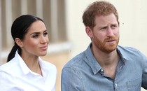 Meghan Markle, wearing a white button-up shirt with her hair slicked back into a low ponytail, and Prince Harry, wearing a chambray shirt, look off to the side of the camera.