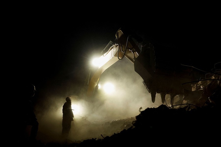 A worker stands beside a large piece of construction equipment, amid dust and rubble.
