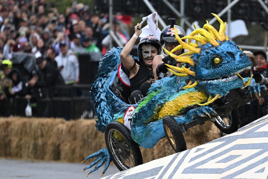 Two people wearing helmets and costumes race in a homemade cart decorated like a large lizard.