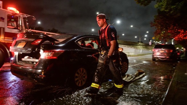 A firefighter walks past a black car parked on the side of a road. A fire truck is parked next to the car and another car is visible further away. 
