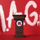 podium with American flags and "MAGA" spray-painted on a red wall in background
