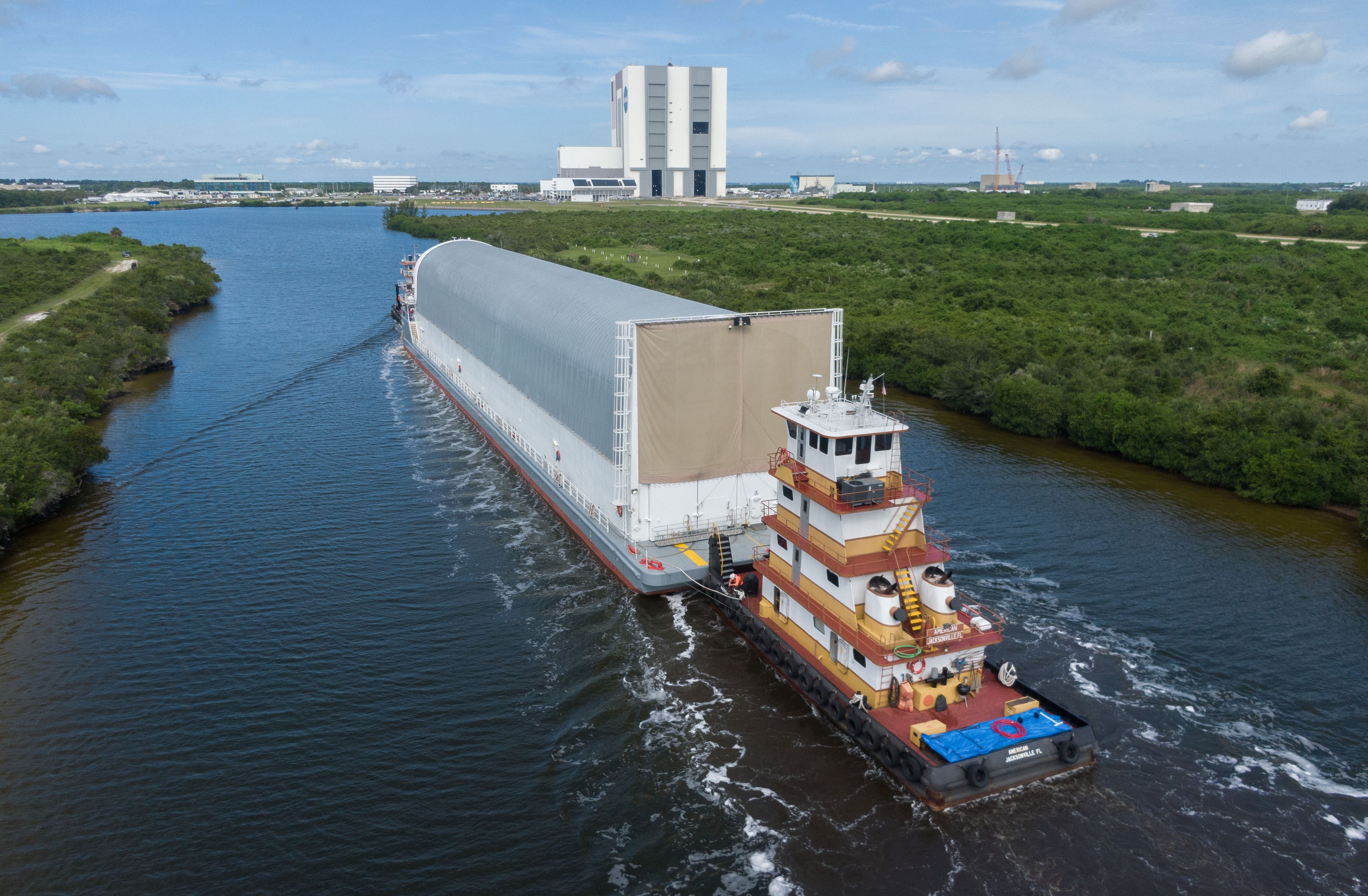Towboats push a long floating container along a channel toward a large rocket launch facility.