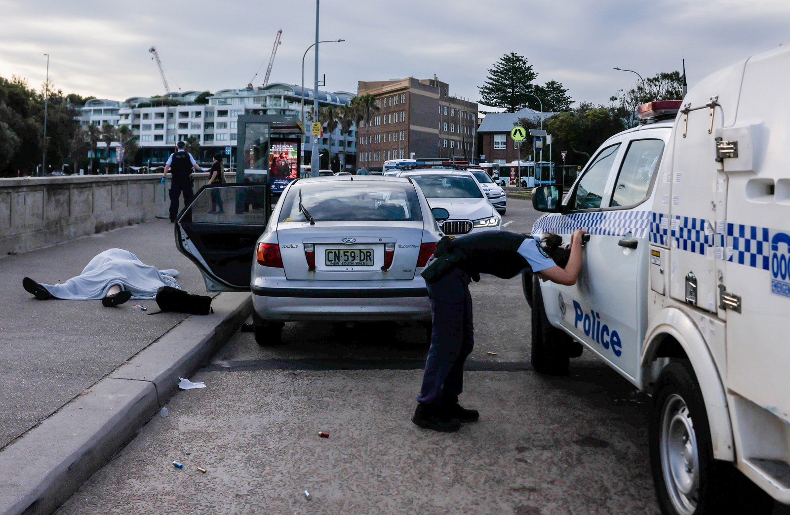 A police officer leans against a police vehicle at the scene of a terrorist attack, with two bodies, covered by a sheet, lying on a sidewalk nearby.