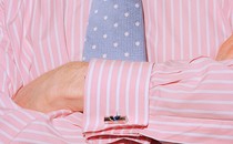 Close-up of a torso of a man wearing a pink dress shirt with cuff links and a blue tie, his arms crossed.