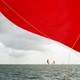 Boats with bright red sails on the ocean, one sail close up to the camera and covering the top half of the frame