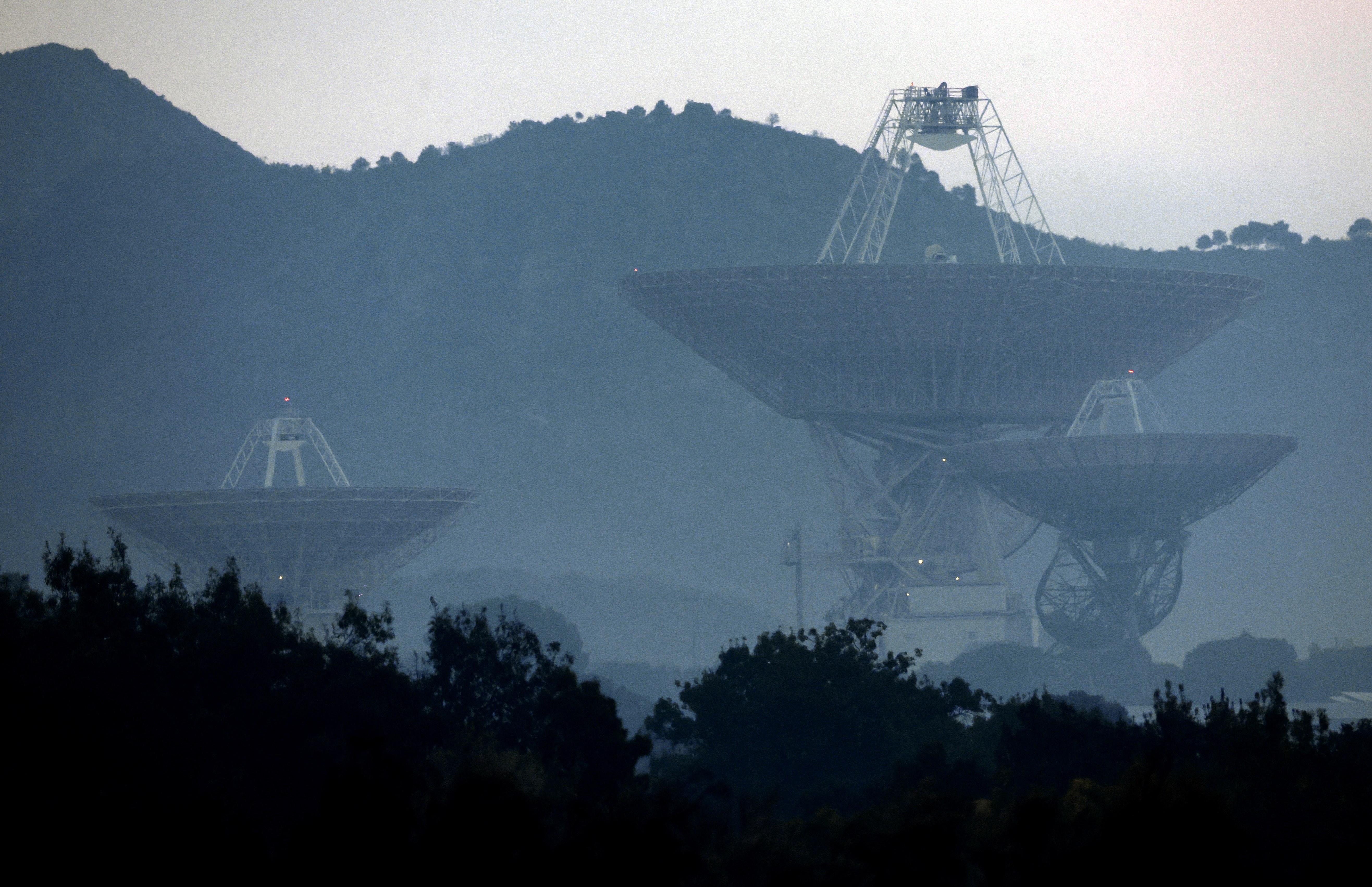 An array of several large radio antenna dishes, seen among hills under smoky skies