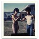 A polaroid of two people facing the camera, wearing casual clothing and afros, with their palms clasped together.