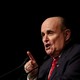A close-up of Rudy Giuliani speaking on a stage in front of a black backdrop