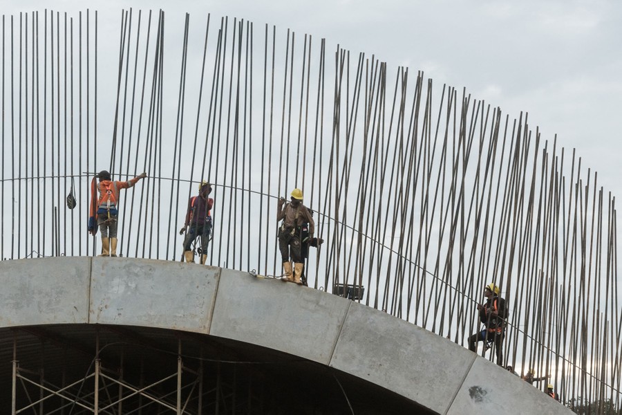 Workers stand along the top of an arched concrete structure being built, beside many reinforcing steel bars.
