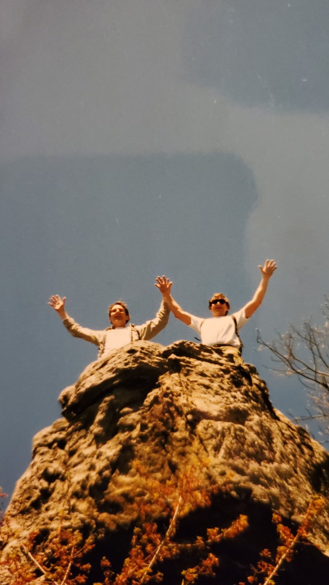 Two men raising their arms in celebration, seen from below as they stand atop a giant rock cliff
