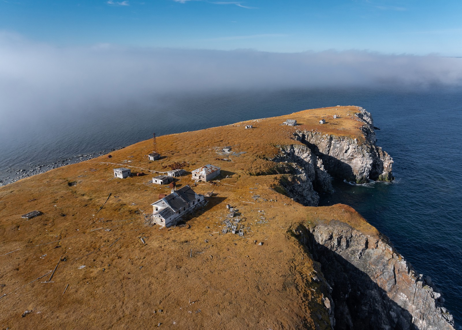 An aerial view of a cluster of small abandoned buildings on a treeless island with a steep cliff shoreline