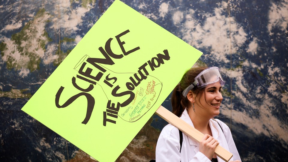 A female protester in a lab coat holds a sign saying: 'Science Is the Solution'