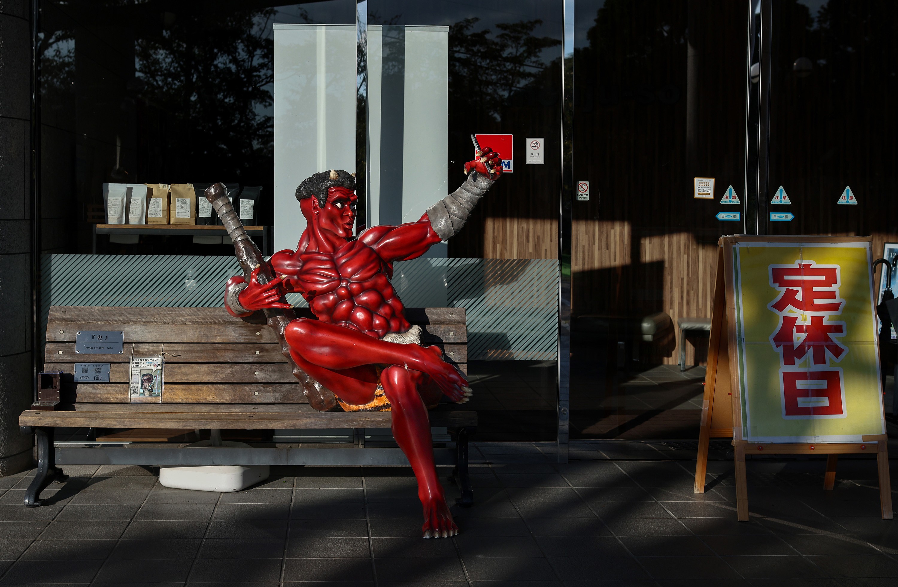 A statue of a demon taking a selfie sits on a bench in front of a storefront.