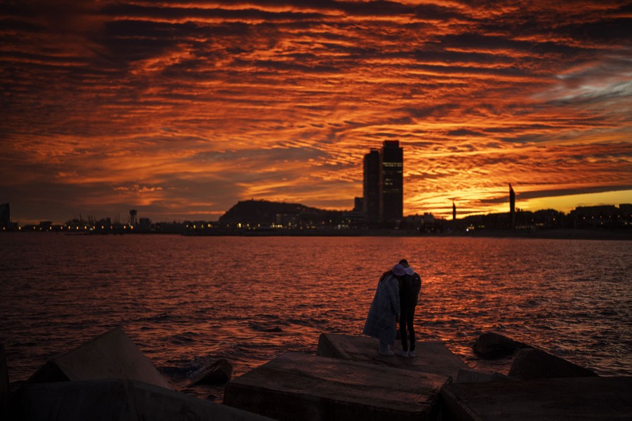 A couple stands near a body of water, watching a colorful sky as the sun sets.