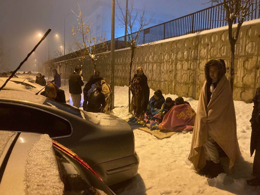People stand in snow, wearing blankets, near cars in a parking lot.