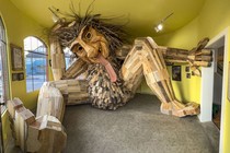 A large wooden troll sculpture sits inside a room, looking cramped, its head against the ceiling.