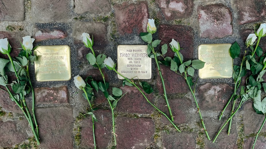 Closeup of a brick street with gold-colored metal blocks inlaid. Left to right they read: Arthur Kahn, Fanny Weinberg, Nathan Weinberg. All three are surrounded by white roses.