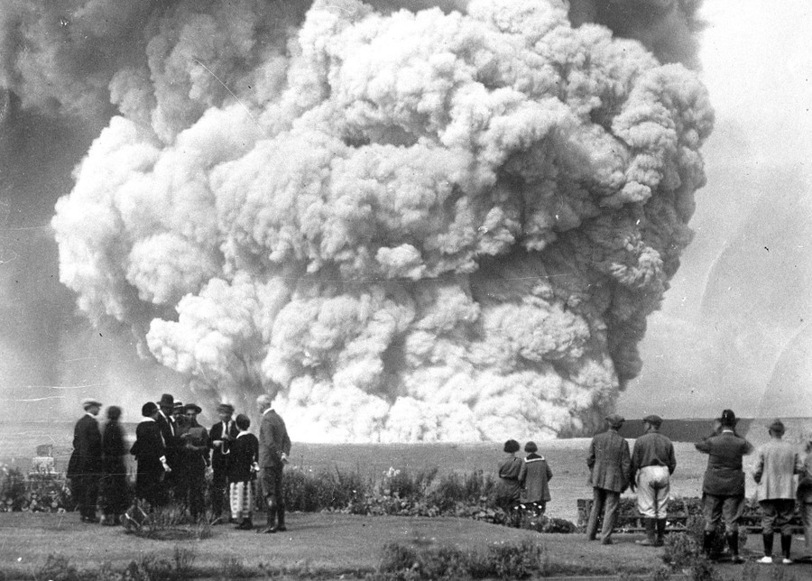 Small crowds of people stand at an observation area, looking toward a huge volcanic eruption in the backround.