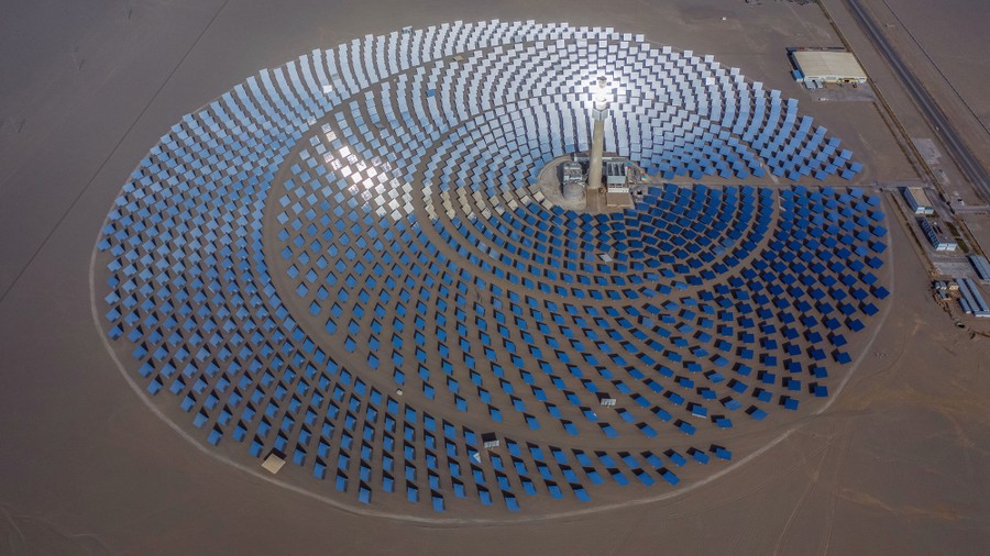 An aerial view of a solar power plant made up of thousands of mirrors in a desert, aiming sunlight at a central tower