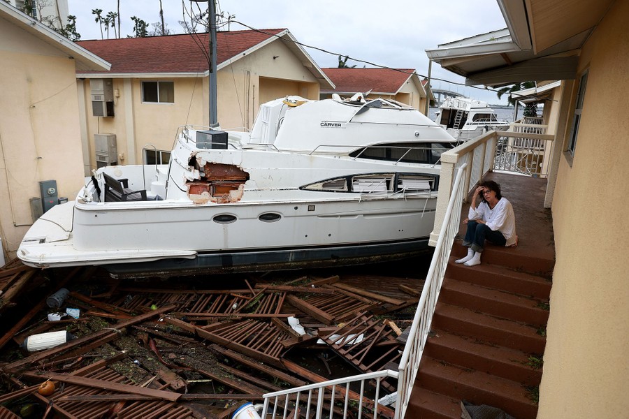 A person sits on steps overlooking a parking lot filled with storm debris and a couple of large boats.
