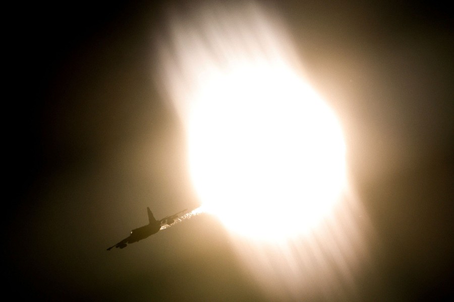 A jet flies through the sky, with a hazy sun seen in the background.