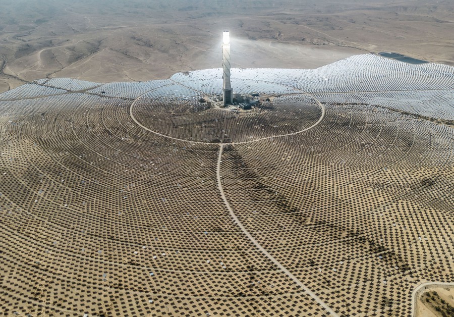 An aerial view of a solar-power station, with thousands of mirrors surrounding a tall tower in a desert