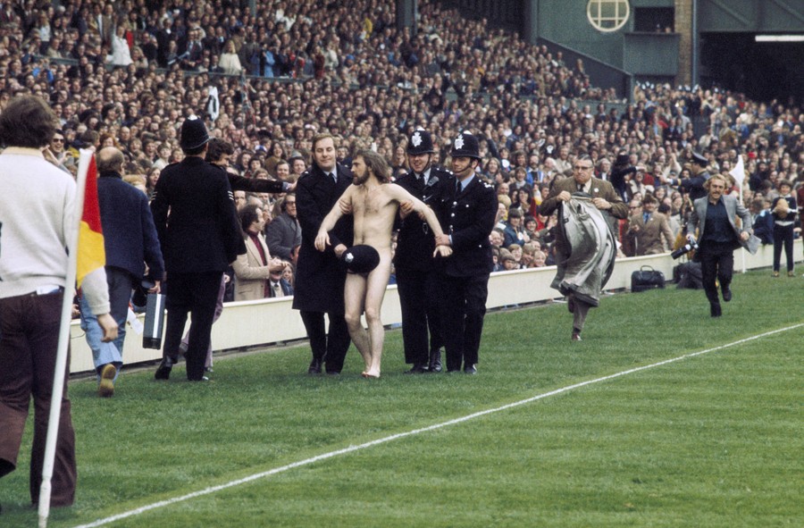 Several police officers escort a naked man off of a rugby field in front of a large crowd.