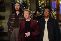 Three mean stand on a stage in front of a set decorated with wreaths and Christmas trees.