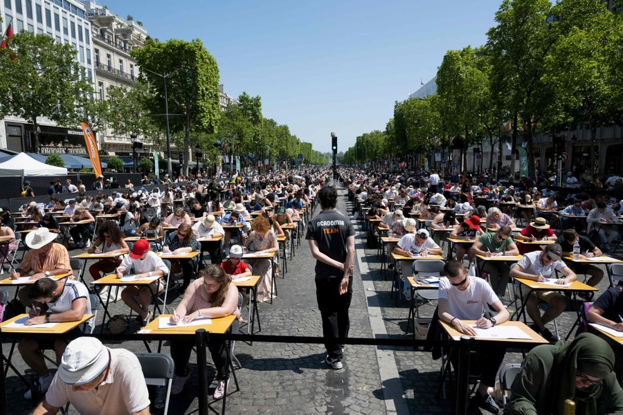 Hundreds of people sit at desks set up in long rows in a wide avenue.
