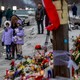 Two little girls in purple coats (one holding a stuffed animal) stand near a flower memorial. Other people are seen in the background.