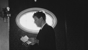Black-and-white photograph of John Hollander reading from loose folded pages