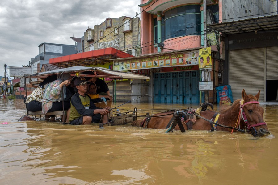 People ride on a horse-drawn cart through deep water in a flooded street.