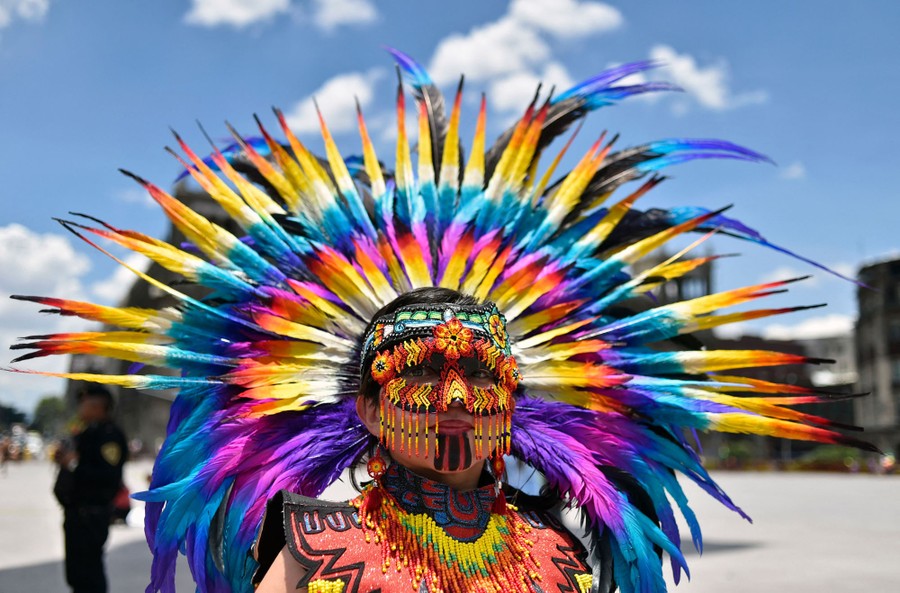 A person performs in a city square wearing a colorful mask and feathered headdress.