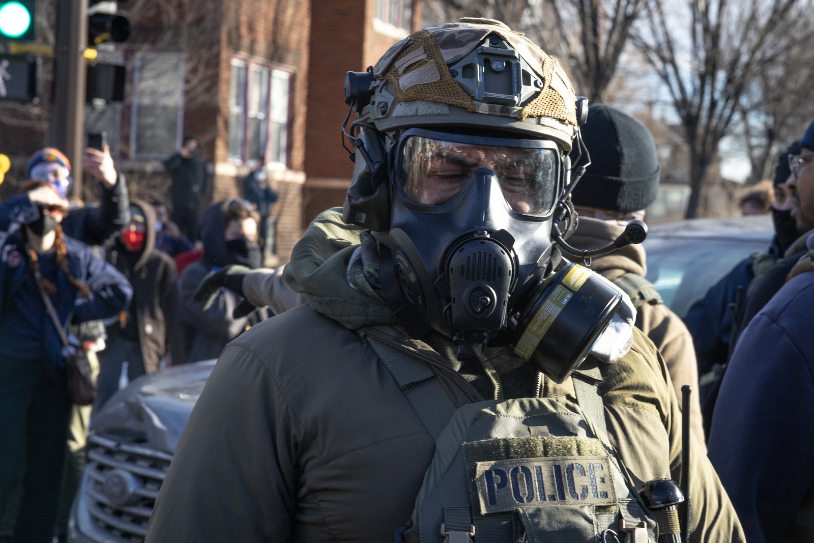 A federal agent wearing a gas mask and helmet is seen in the foreground with onlookers and protesters visible in the background.