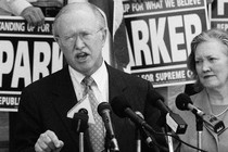 A black-and-white photo of a man speaking at a lectern with microphones. Behind him, people hold signs that read "Standing Up for What We Believe: Parker, Republican for Supreme Court."