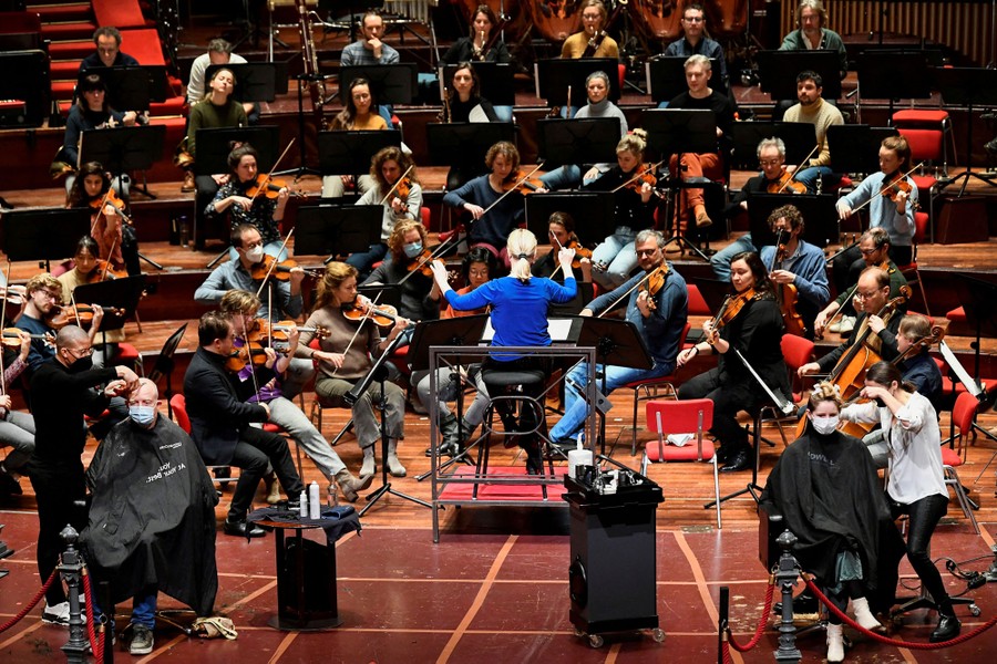 Two people get their hair cut on a stage as an orchestra plays behind them.