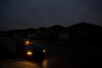 A night view of a people in a car with its interior lights and headlights on, parked in a suburban street surrounded by dark houses.