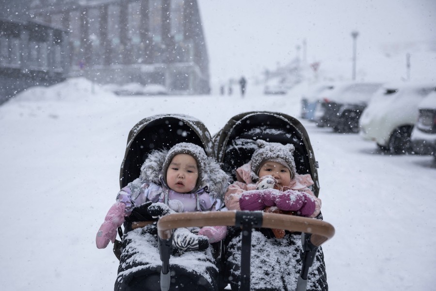 Two young children, bundled up, sit in a two-seat stroller in a snow-covered parking lot.