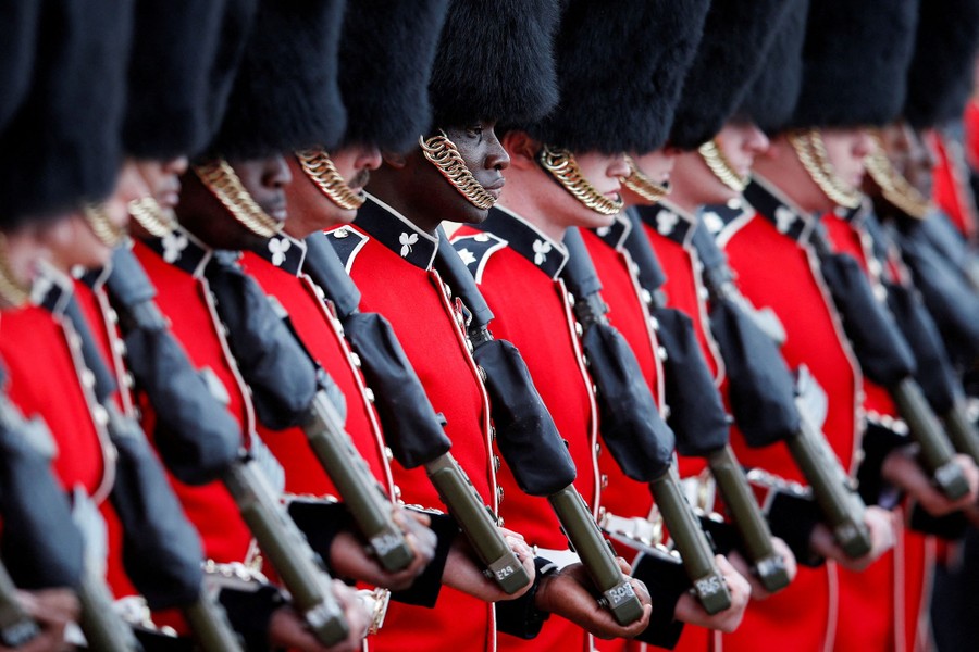 Soldiers wearing red uniforms and tall, black, fuzzy hats stand at attention.