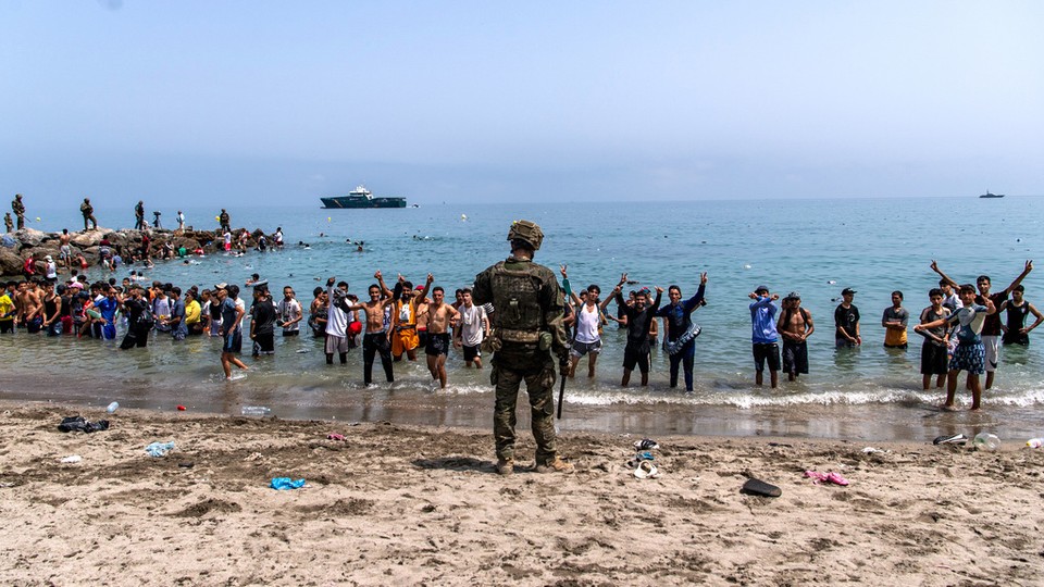 A solider stands on a beach littered with trash and filled with people approaching the shore from the water.