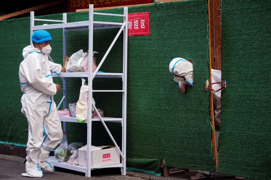 A worker in a protective suit looks through an opening in a barrier toward another worker standing beside a rack with several bags on it.