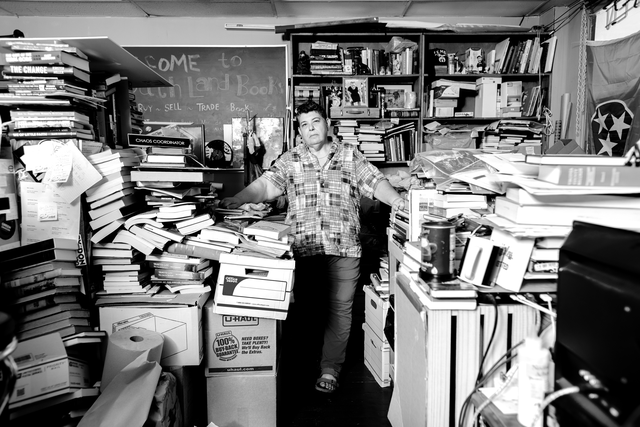 black-and-white photo of woman in plaid shirt and jeans standing in middle of crowded bookstore among piles and shelves of books