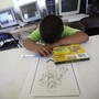 A child rests his head on the table next to a box of markers and a coloring page.