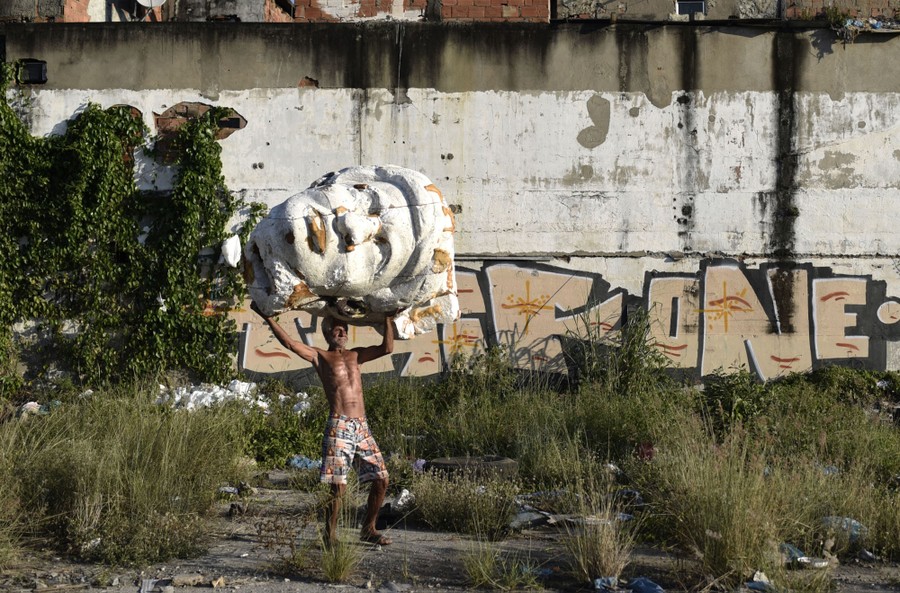 A person carries a large crumbling sculpture of a head through a vacant lot.